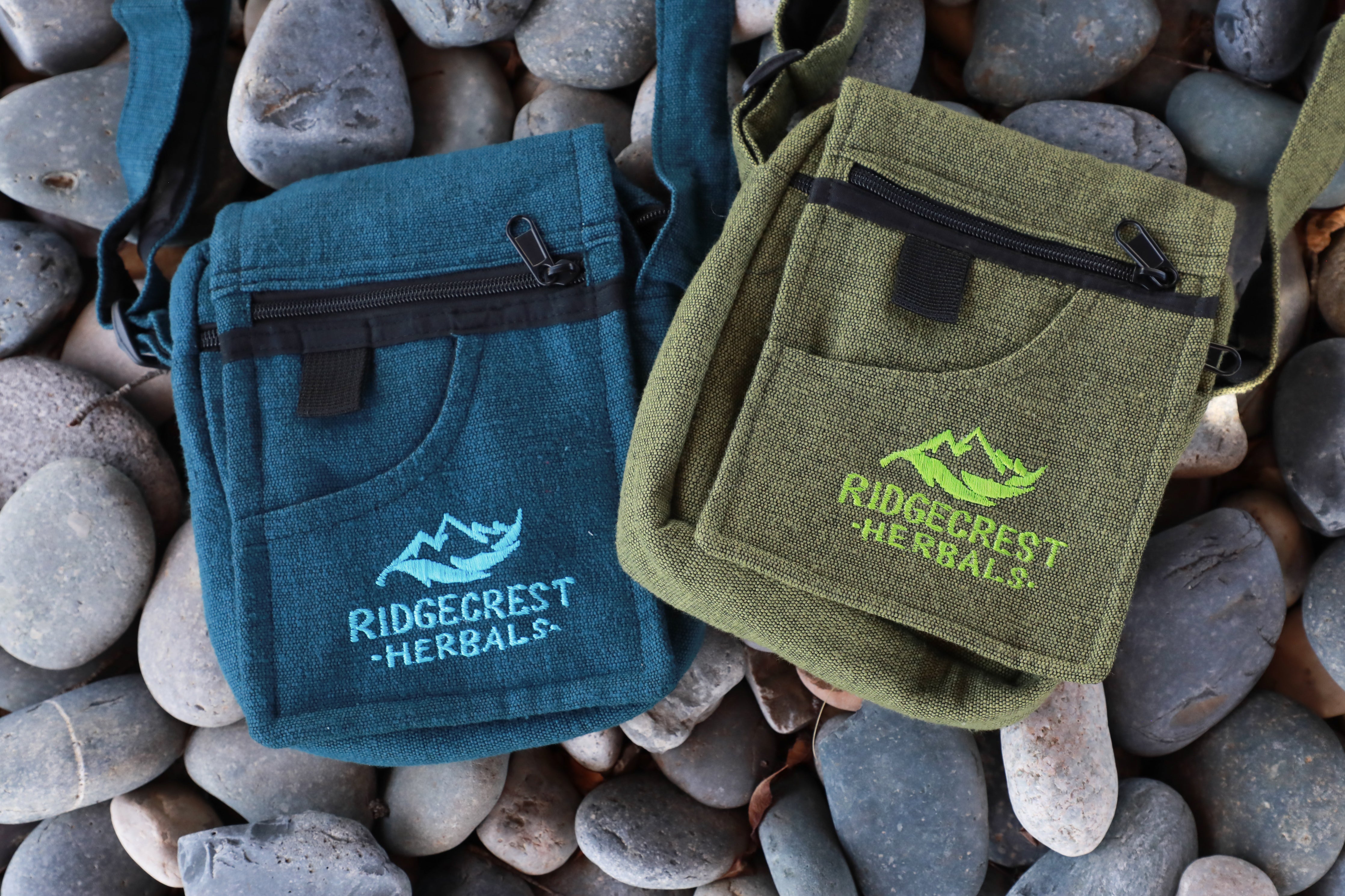 Two Ridgecrest Herbals adventure satchels, one green and one blue on a background of pebbles