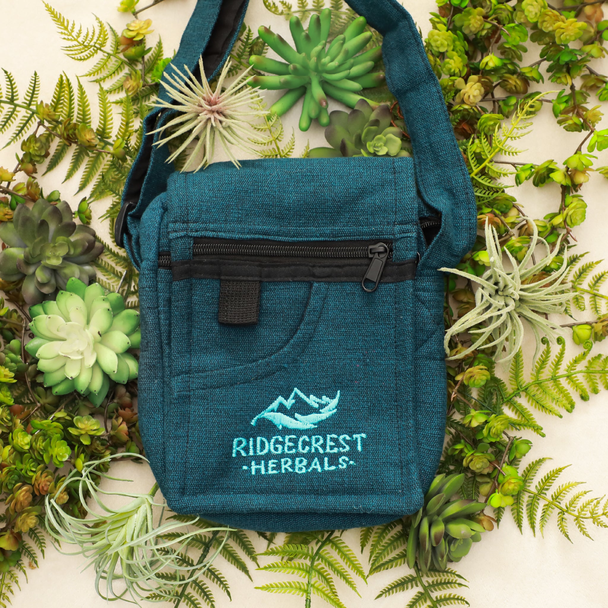 A blue Ridgecrest Herbals adventure satchel laid on green ferns and succulent plants