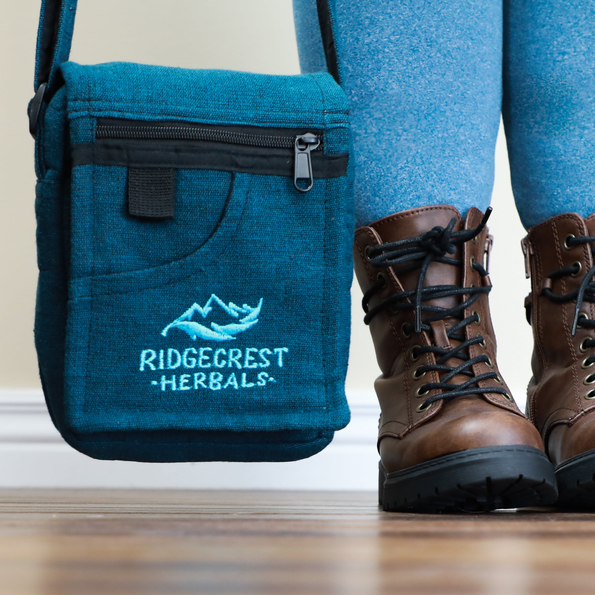 A blue Ridgecrest Herbals adventure satchel hanging near a person wearing brown hiking boots and blue leggings, standing on a wood floor.