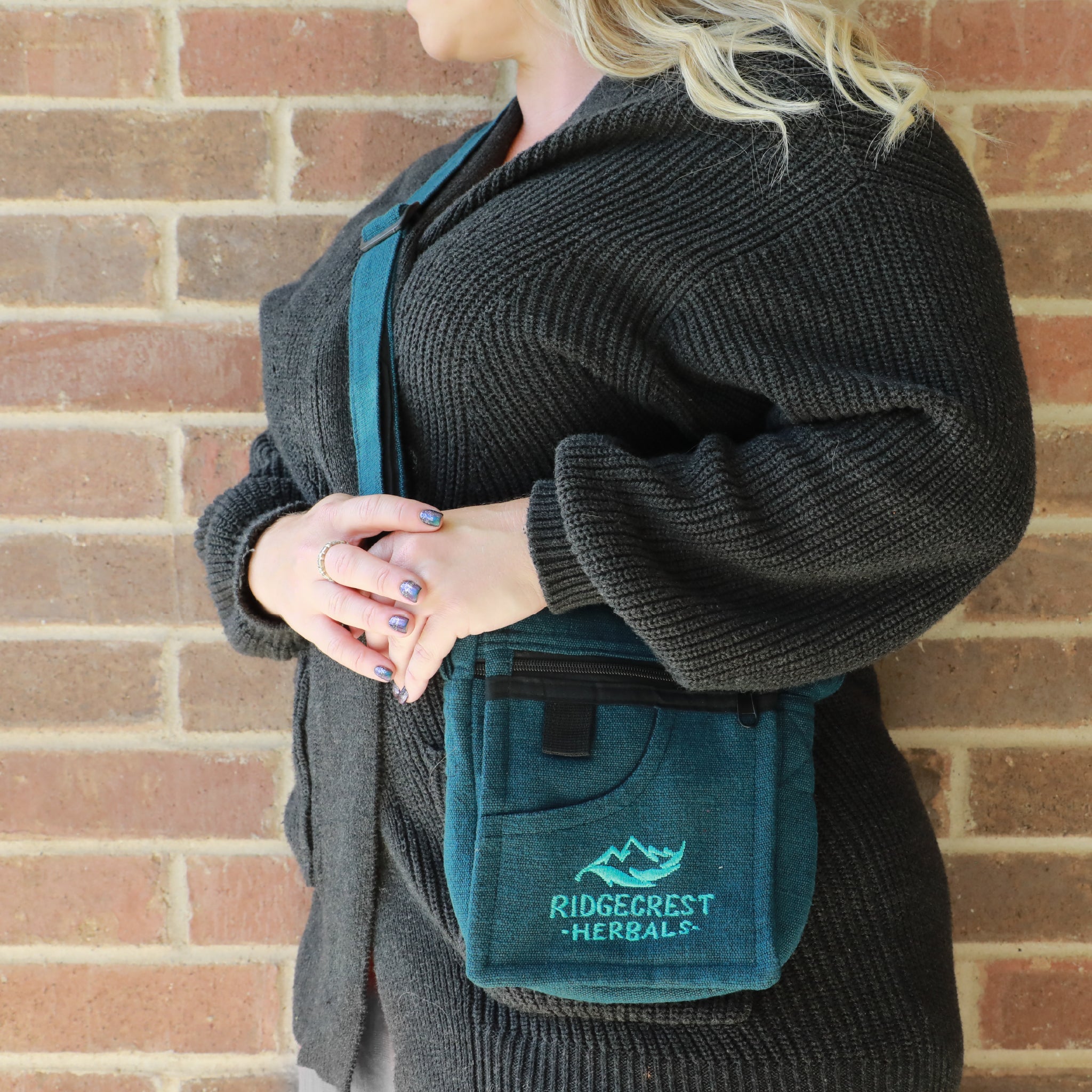 A woman wears a blue Ridgecrest Herbals adventure satchel standing against a brick wall.