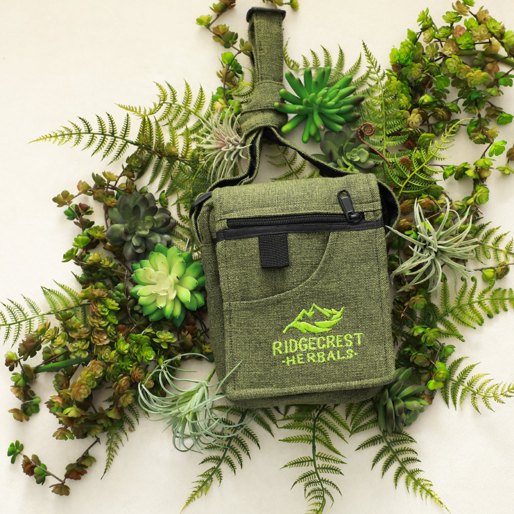 A green Ridgecrest Herbals adventure satchel laid on green ferns and succulent plants