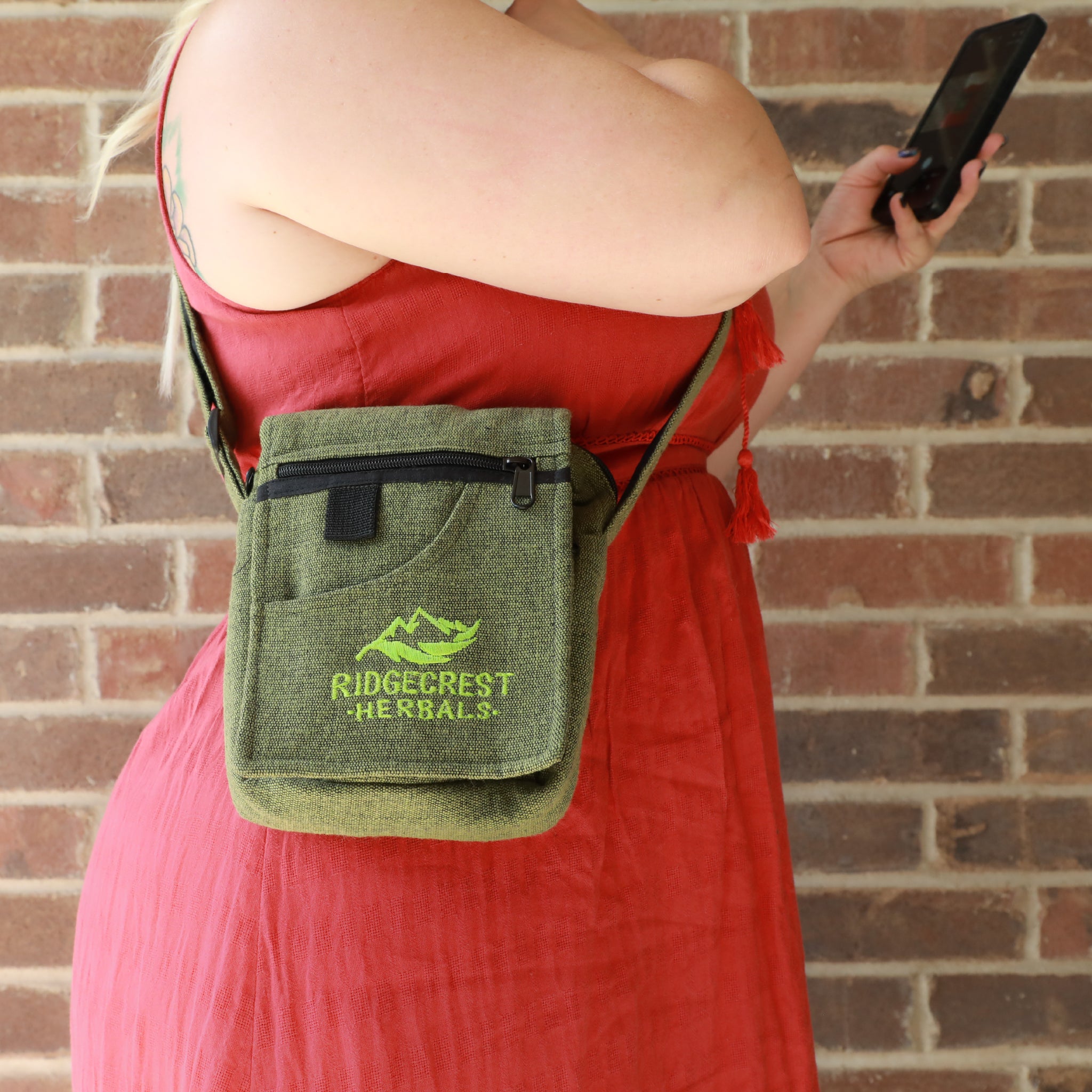 A woman in a red dress wears a green Ridgecrest Herbals adventure satchel standing against a brick wall.