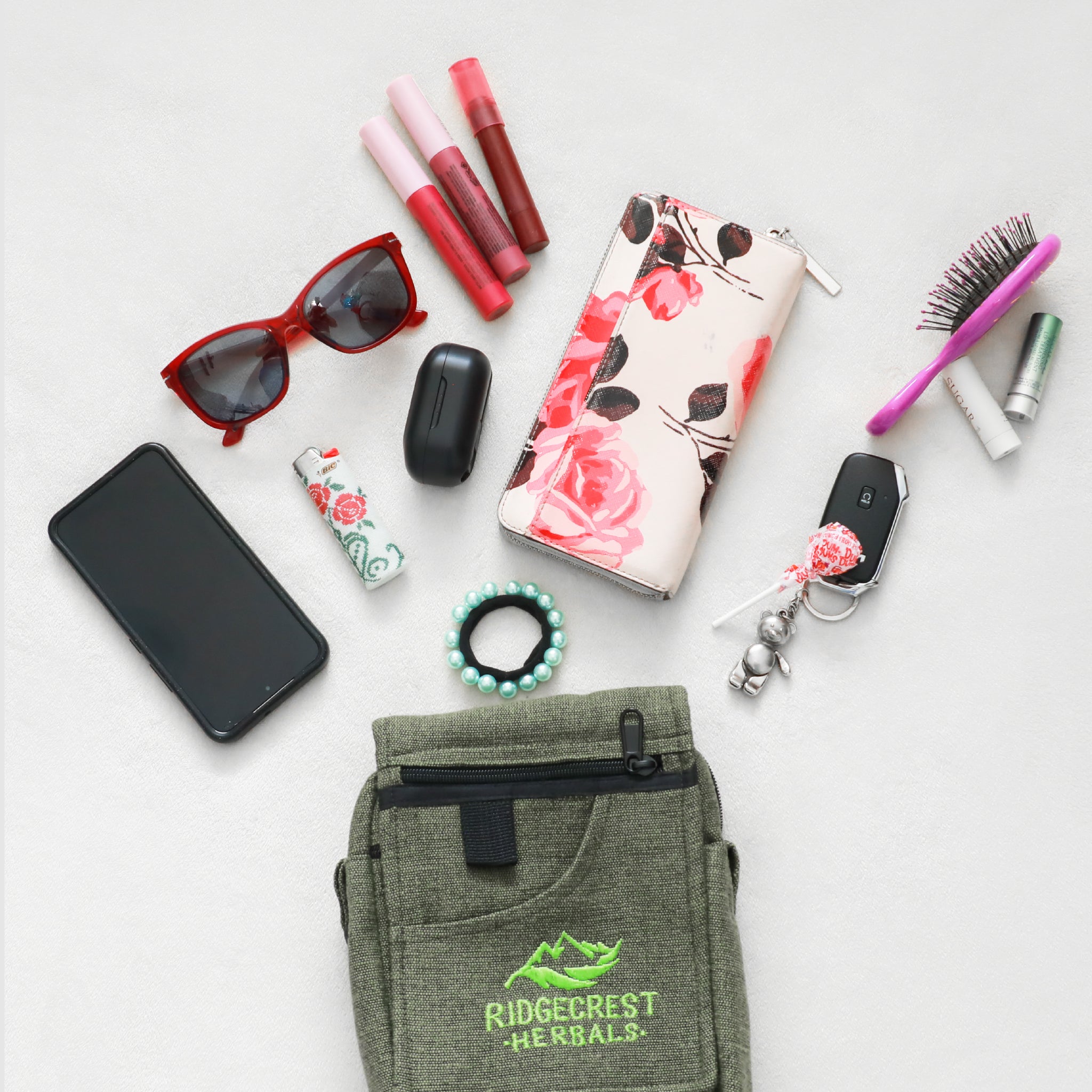 A green Ridgecrest Herbals satchel surrounded by personal items, including sunglasses,a phone on a white background