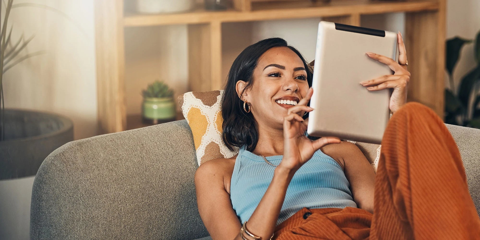 Smiling woman in cozy casual wear relaxing on a couch, using a tablet in a bright, warm-toned living room.