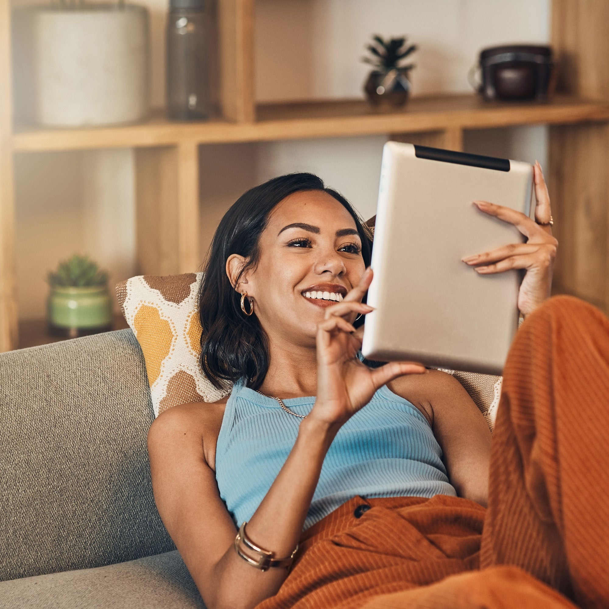 Smiling woman in cozy casual wear relaxing on a couch, using a tablet in a bright, warm-toned living room.