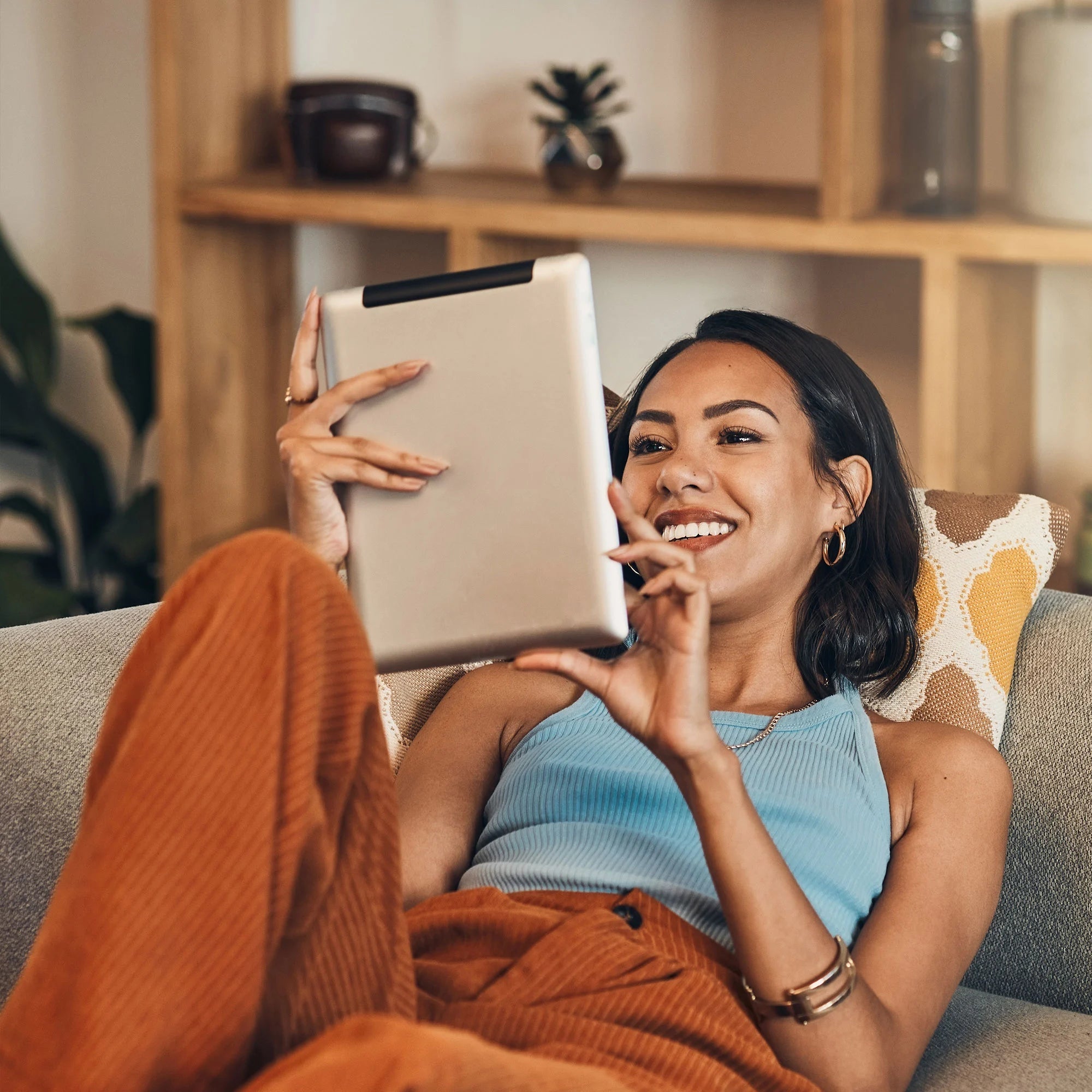 Right-side angle view of a smiling woman in cozy casual wear lounging on a couch, using a tablet in a warm-toned living room.