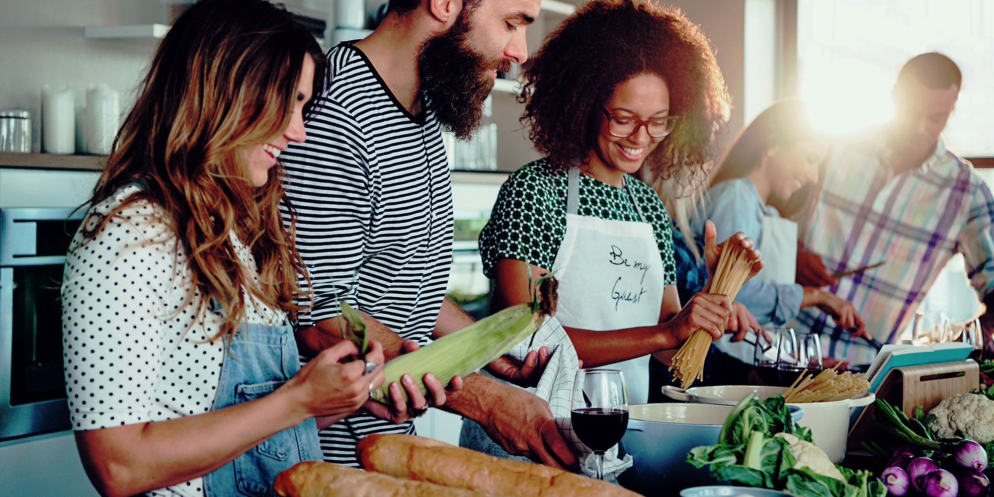A group of smiling friends in a bright kitchen prepare a meal together with fresh vegetables and bread on the counter.