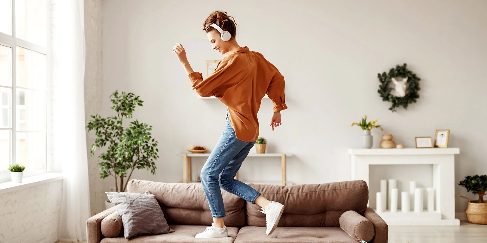 Bright photo of a woman with headphones dancing on a couch in a modern, minimal living room with natural light and cozy decor