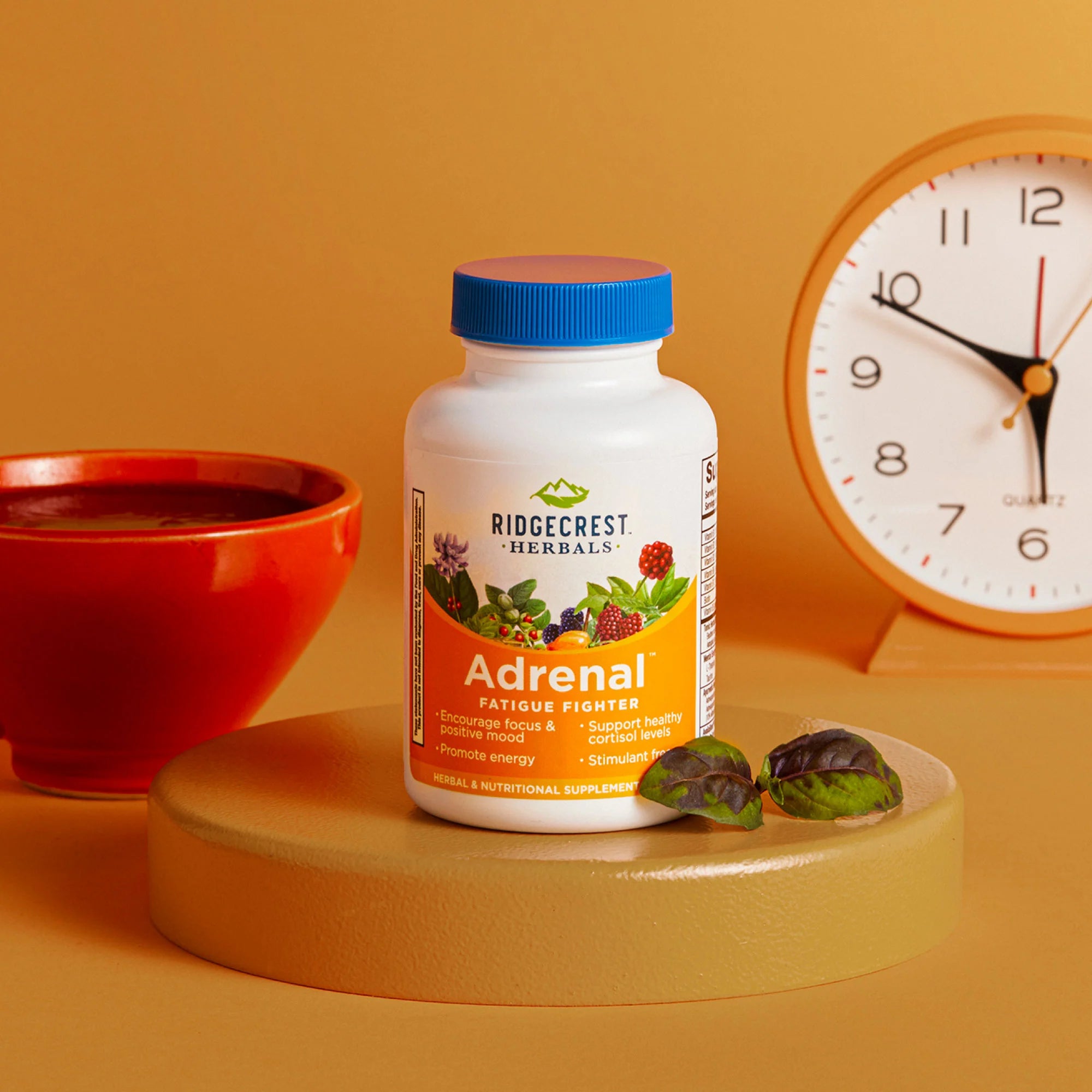 Adrenal Fatigue Fighter supplement is displayed with a red cup, a clock, and fresh basil leaves, set against a warm orange background.