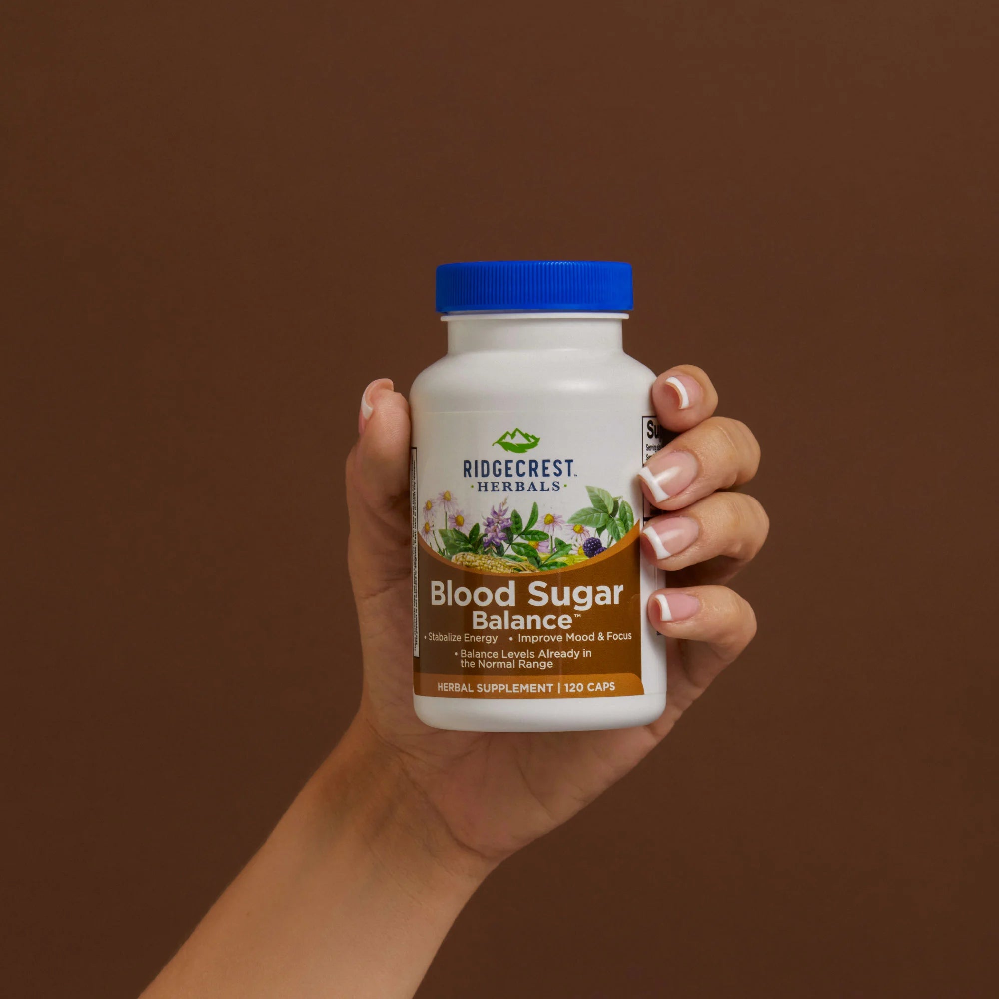 A manicured female hand holds a bottle of Blood Sugar Balance against a brown background