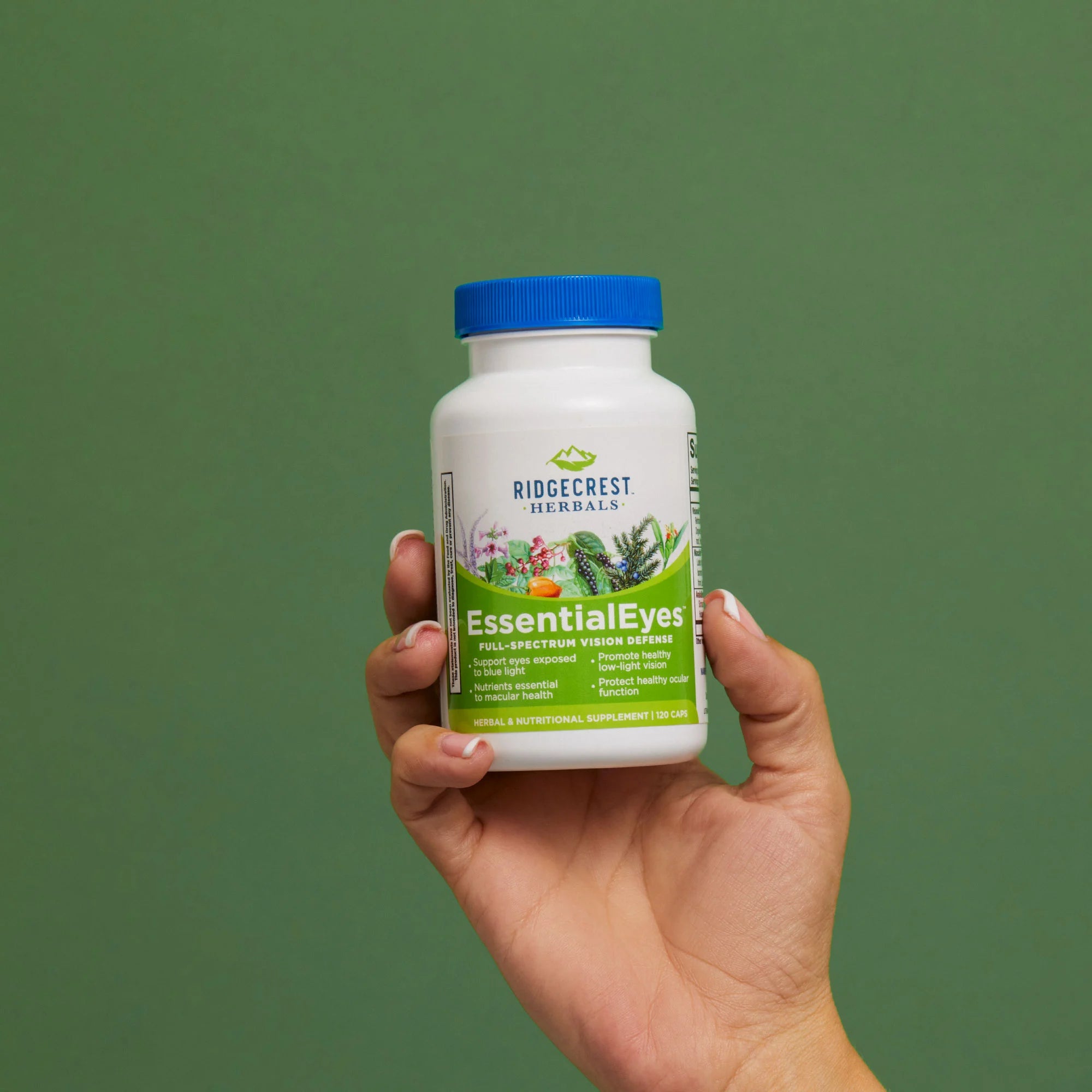 A manicured female hand holds a bottle of Essential Eyes against a green background