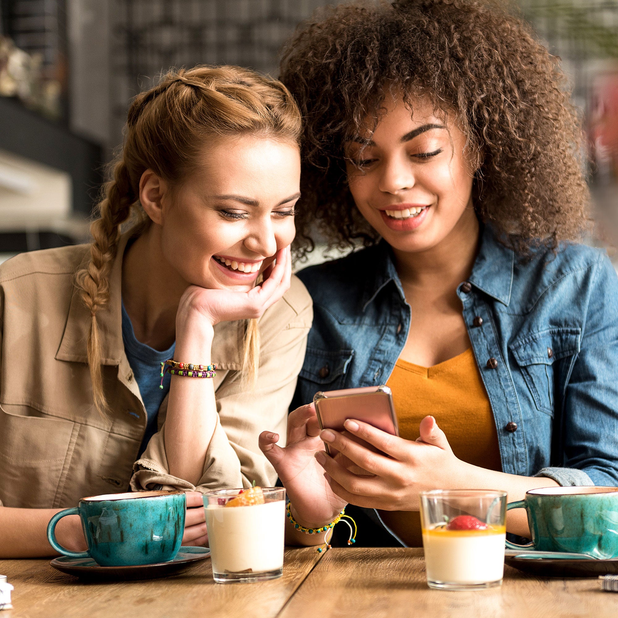 Two smiling women at a café, enjoying coffee and desserts, looking at a phone. One has braided blonde hair, the other has curly hair.