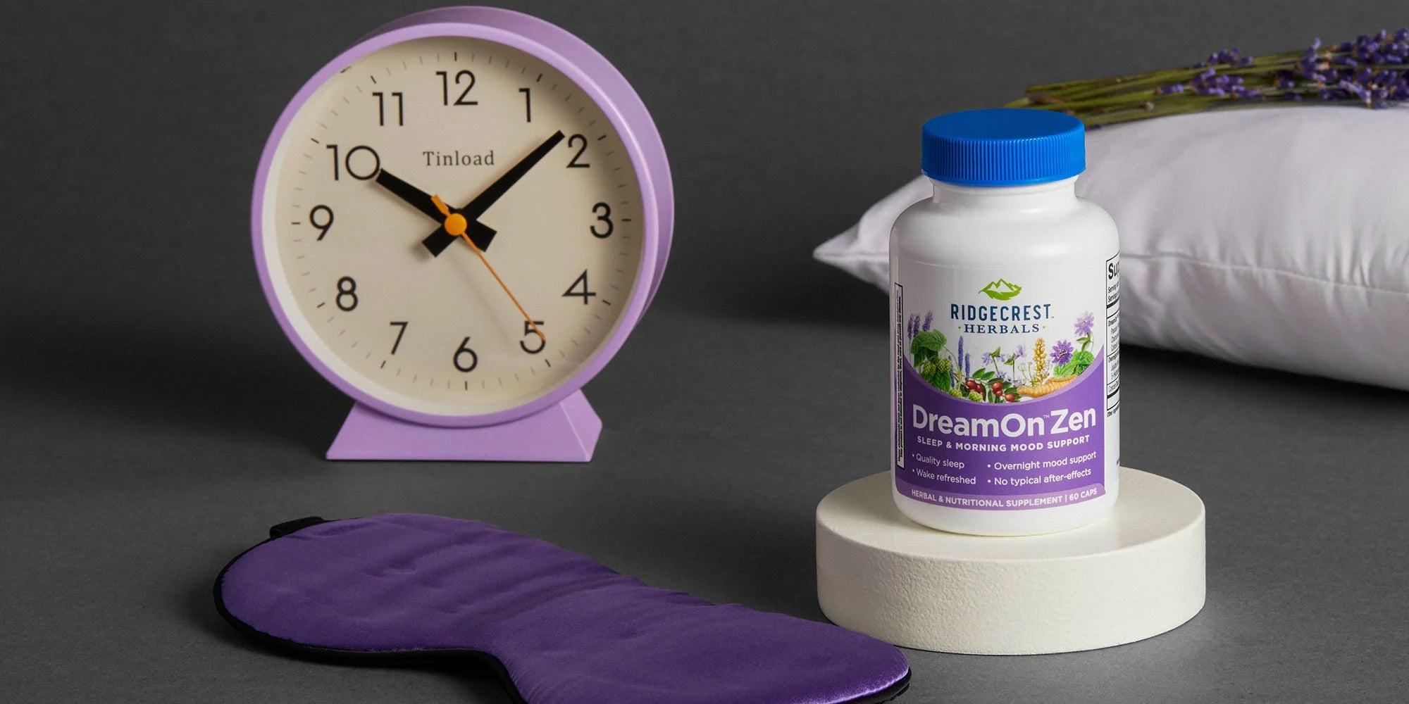 An image with soft purples, whites, and grays. It features a clock, lavender sprigs on a pillow, a purple eye mask, and a DreamOn Zen bottle. 