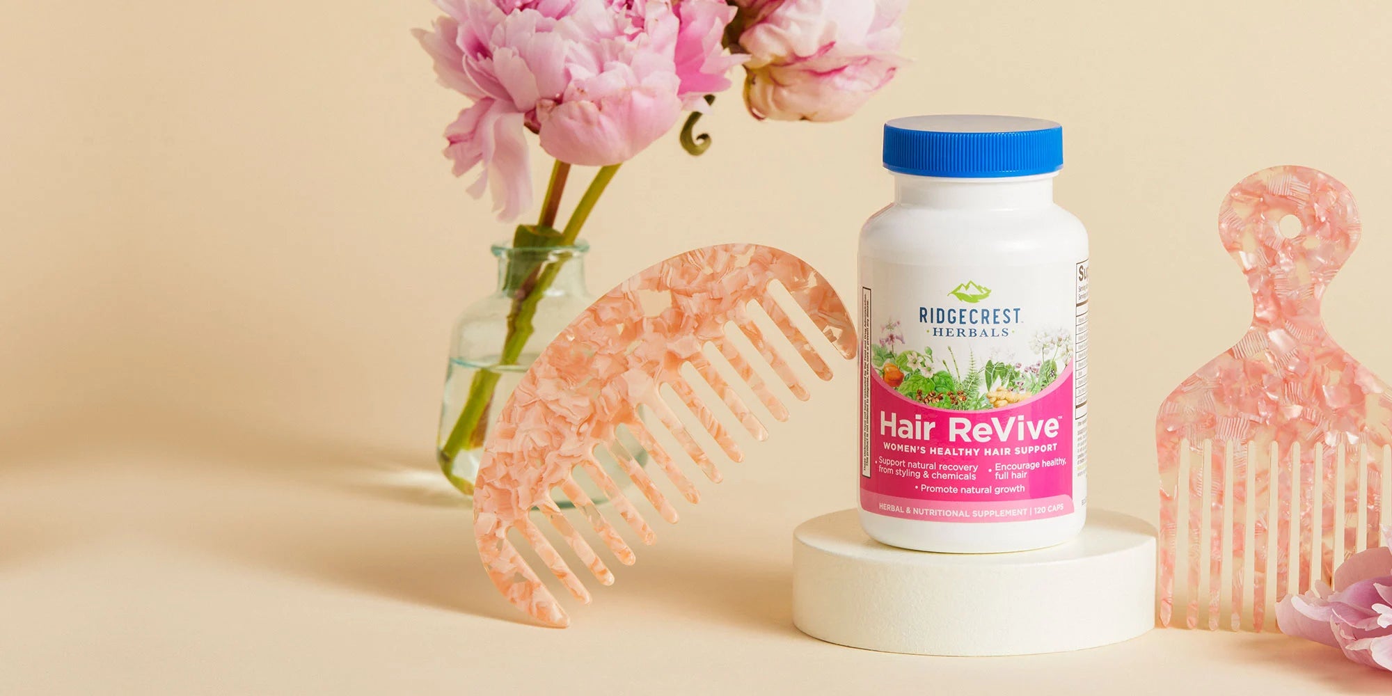 Bottle of HairReVive supplement on cream colored pedestal with pink combs and peonies nearby.