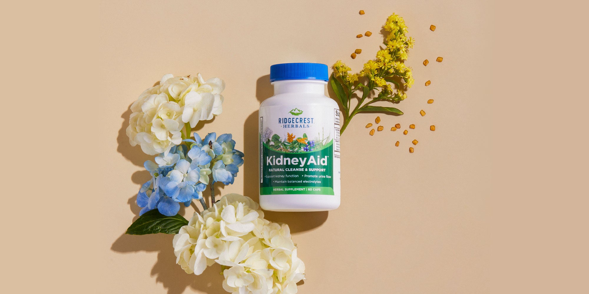 KidneyAid bottle is displayed with white and blue hydrangeas, yellow flowers, and scattered fenugreek seeds on a beige background.