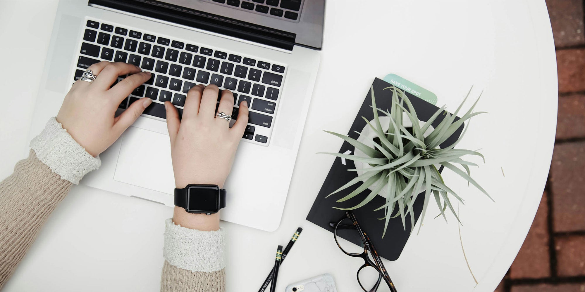 Hands typing on a laptop keyboard, with a nearby plant and notebook on a clean, white desk