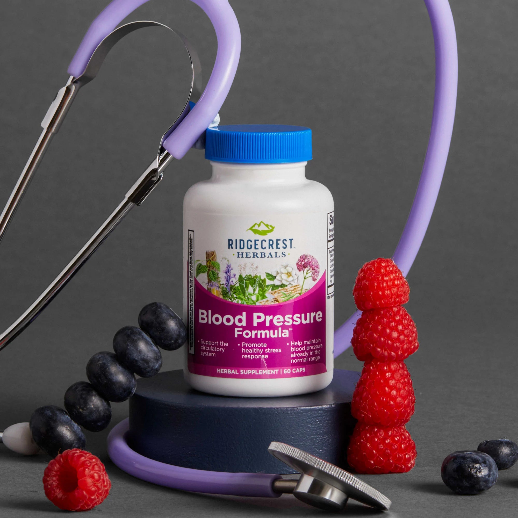 Blood Pressure Formula supplement is displayed with stacked raspberries, blueberries, and a purple stethoscope, set against a dark gray background.