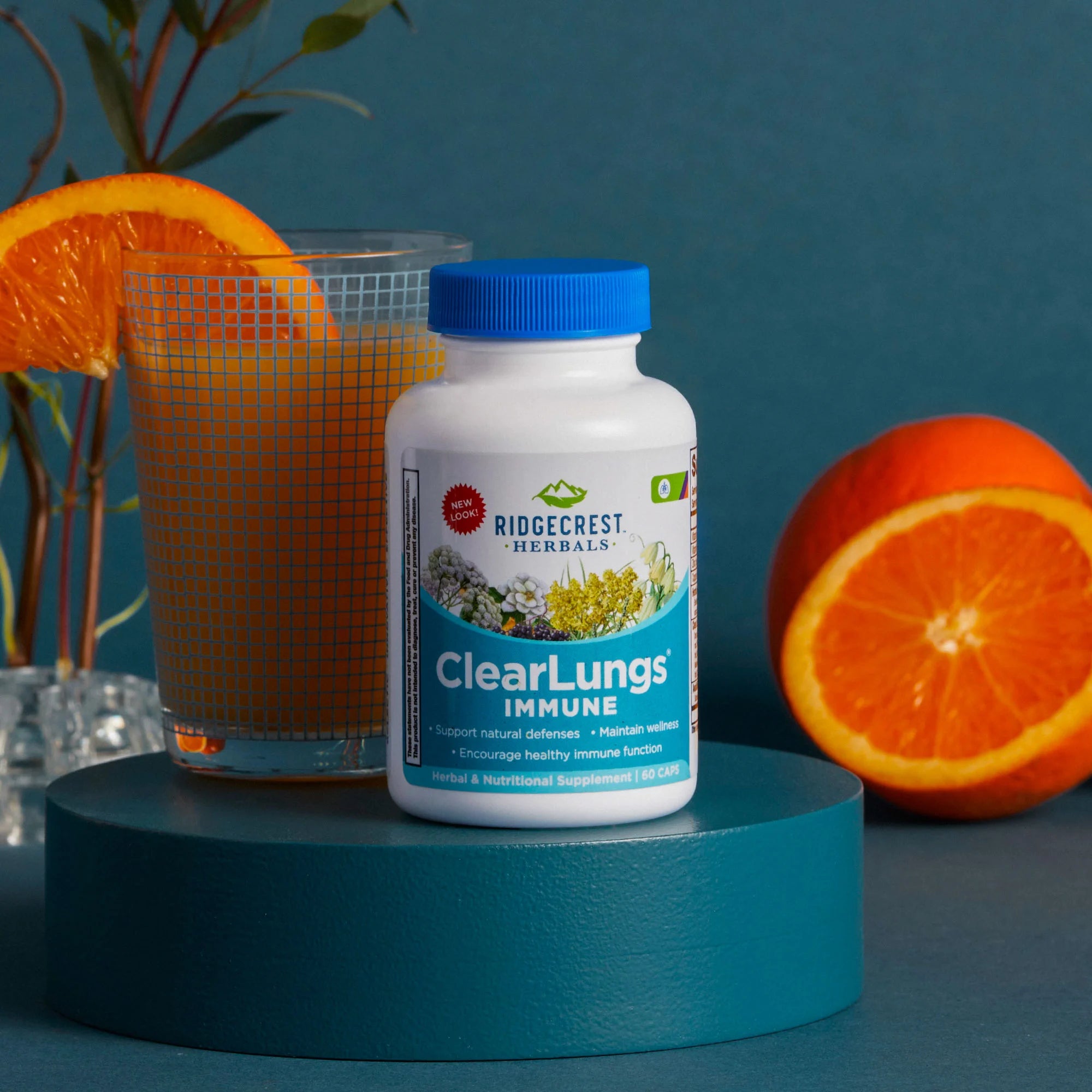 ClearLungs Immune supplement is displayed with a glass of orange juice, a slice of orange, and whole oranges, greenery against a teal background.