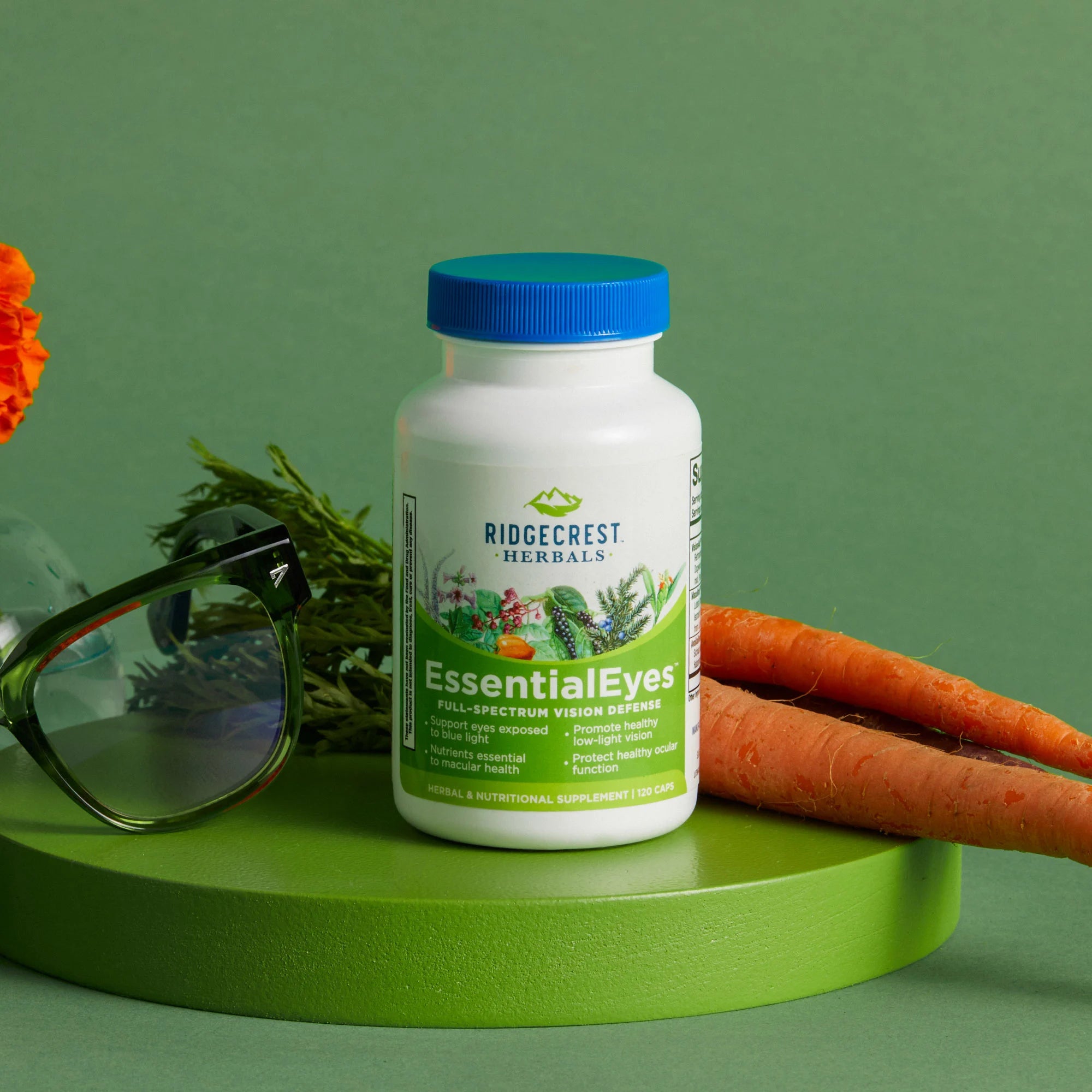 EssentialEyes supplement is displayed alongside fresh carrots, a pair of green-rimmed glasses, and an orange marigold, set against a green background.