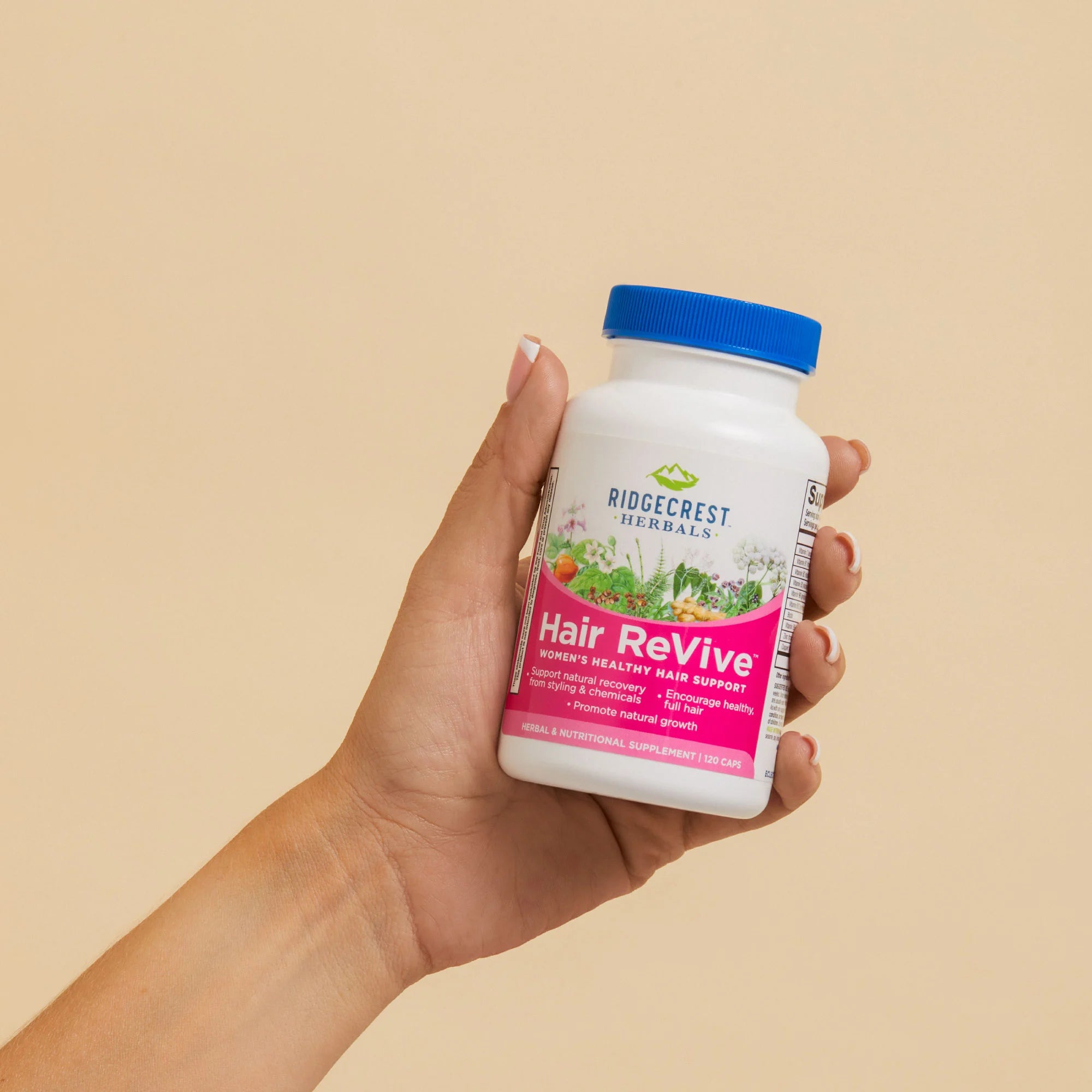 A manicured female hand holds a bottle of HairRevive against a beige background 