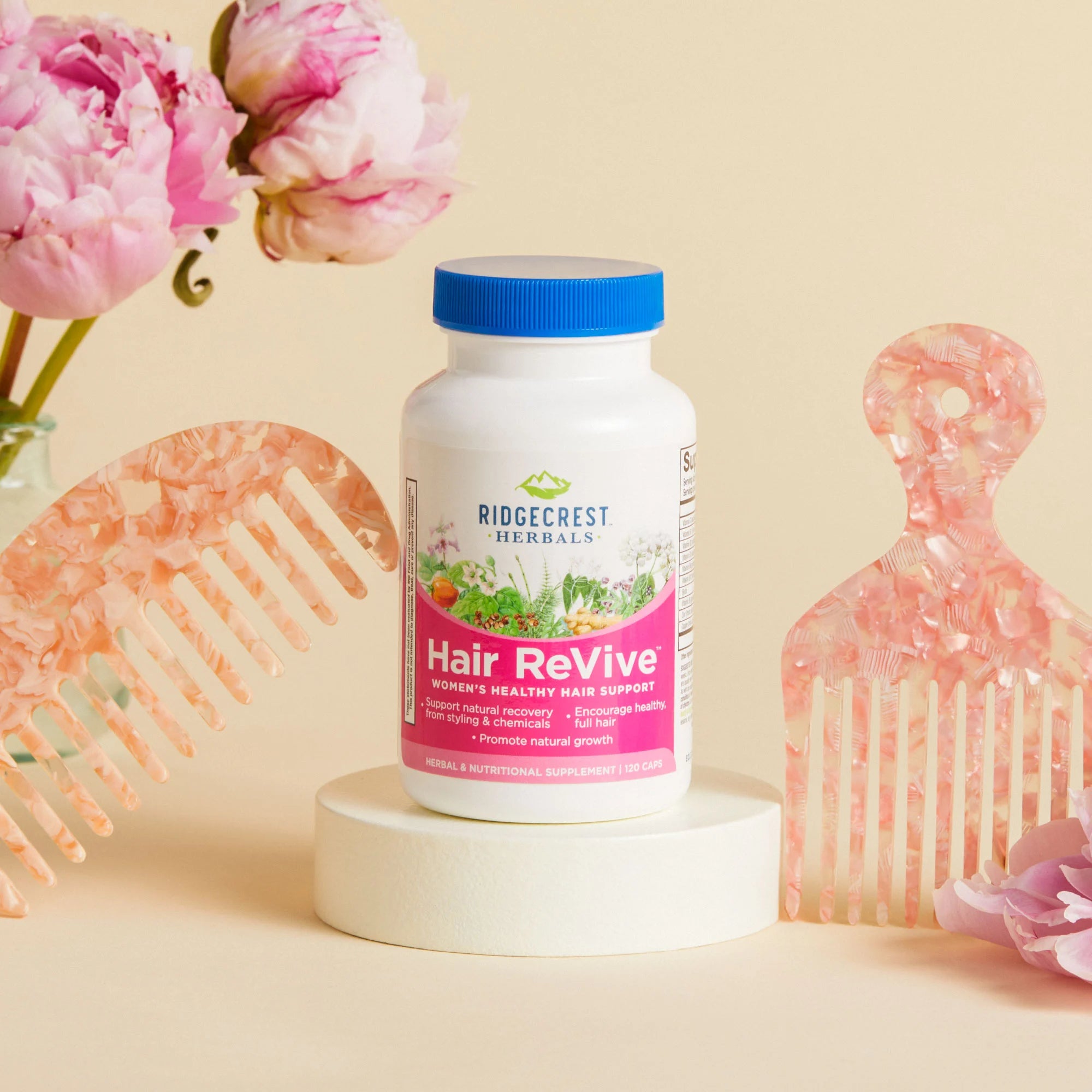 Hair ReVive bottle is displayed with pink peonies and two pink decorative hair combs, set against a soft beige background.