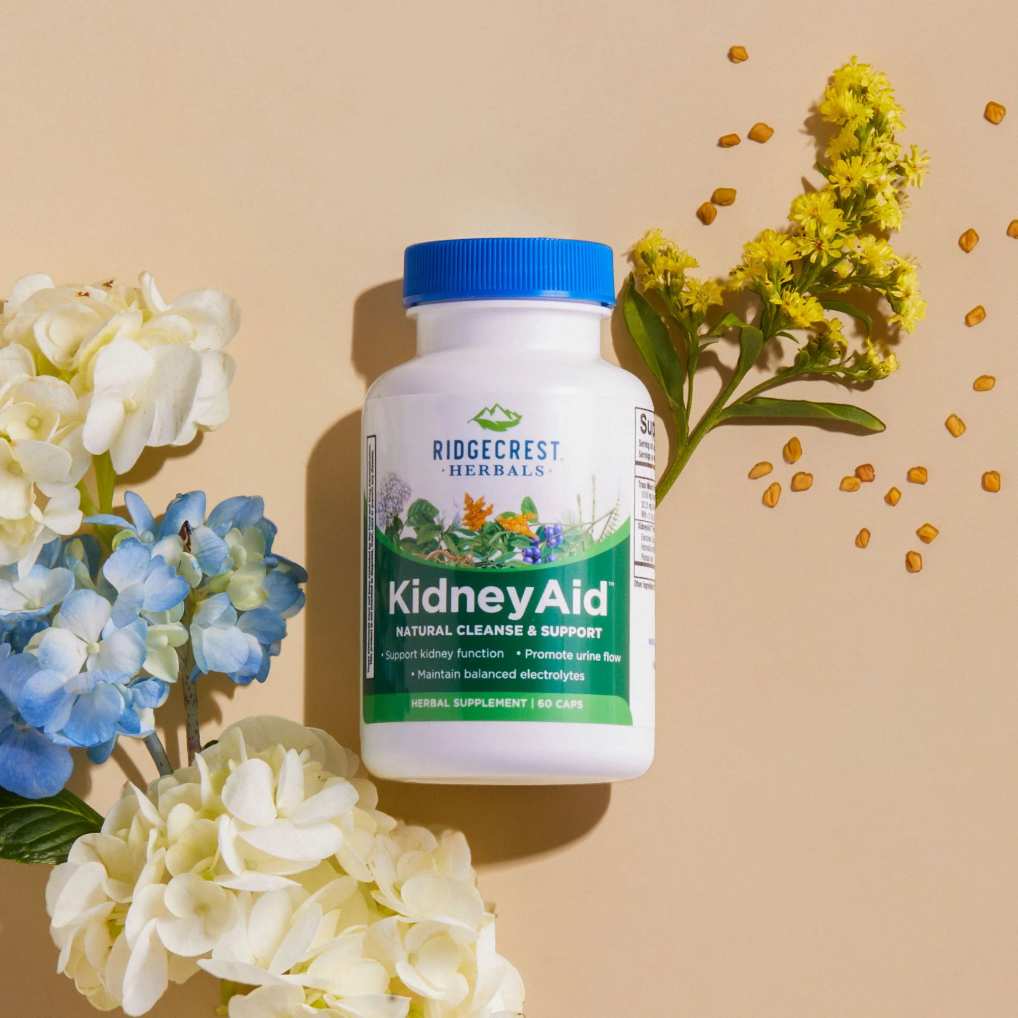 KidneyAid supplement is displayed with white and blue hydrangeas, yellow flowers, and scattered fenugreek seeds on a beige background.