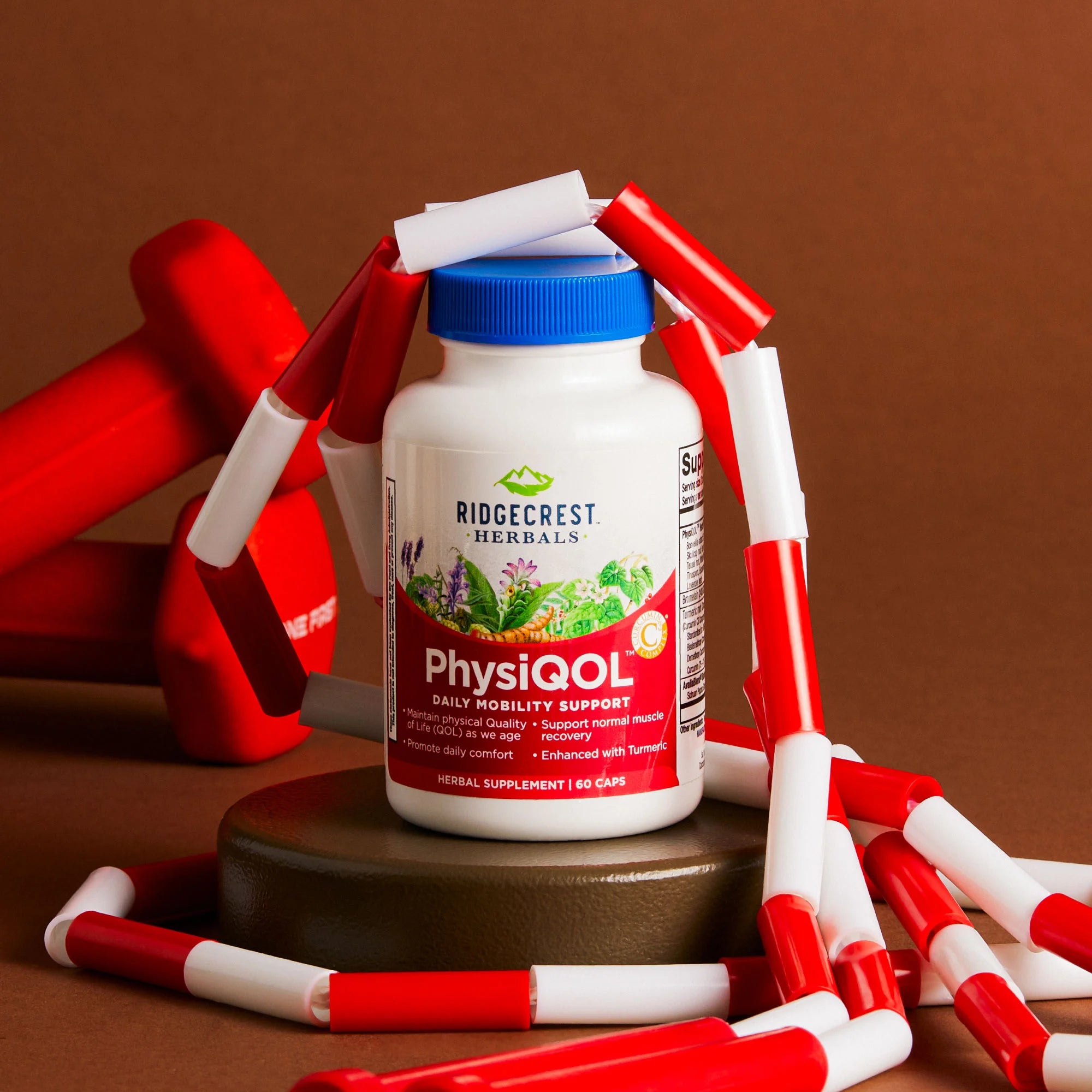 PhysiQOL bottle on a brown round pedestal with red dumbbells and a white and red jump rope, against a matte brown background.