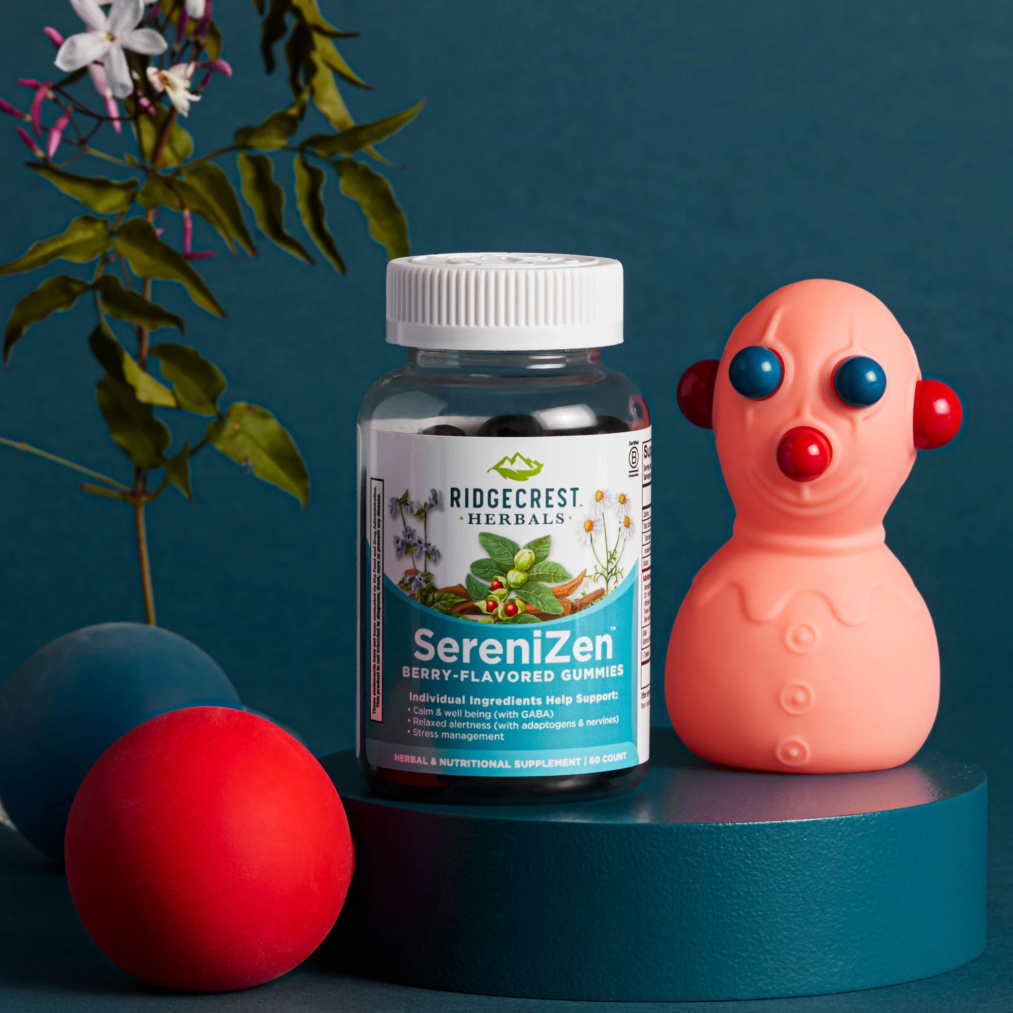 SereniZen Gummies bottle on a teal pedestal with a toy figure, colorful balls, and a flowered stem against a dark teal background