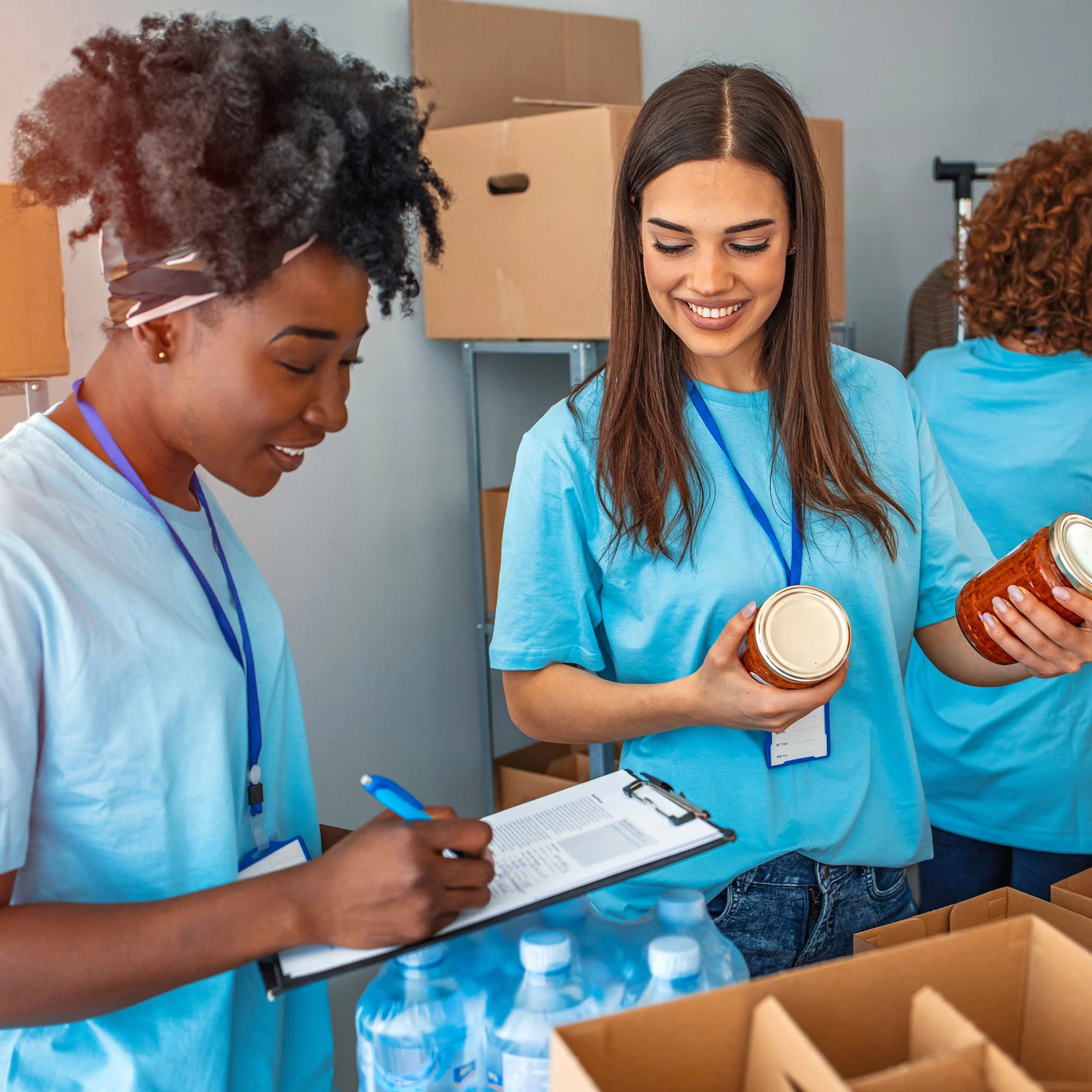 Two women volunteering, organizing donations. One holds canned goods while the other writes notes on a clipboard.